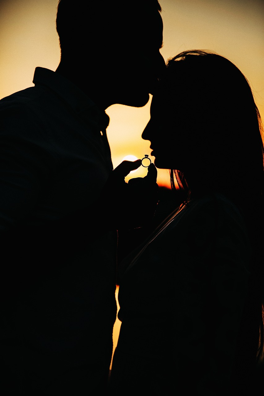 Silhouetted couple at sunset holding an engagement ring during a romantic proposal moment.