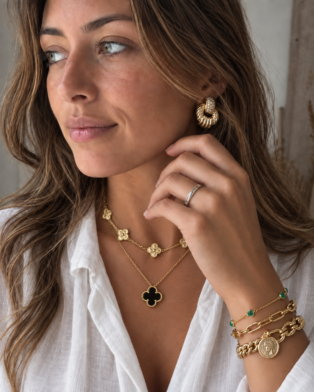 Woman wearing layered gold necklaces, statement earrings, and stacked bracelets with emerald and coin details.